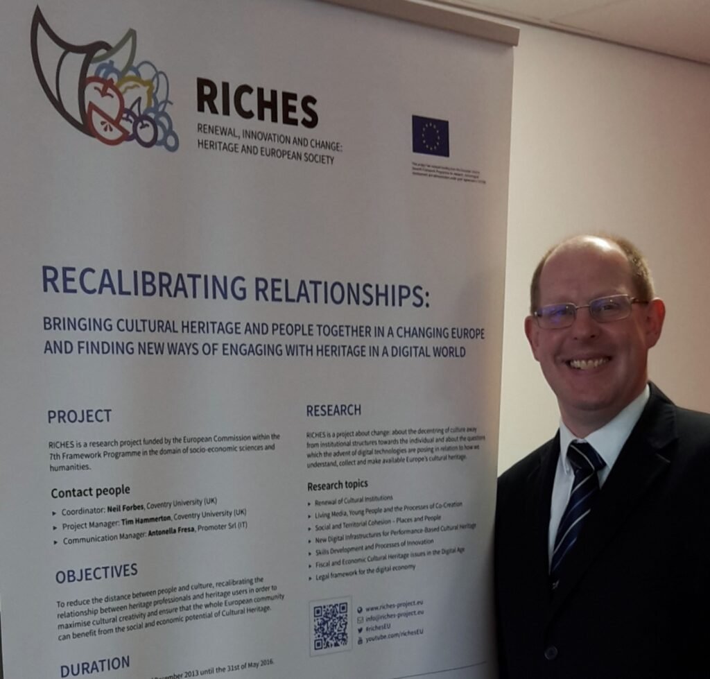 Tim Hammerton, in suit and tie, standing next to the RICES project banner that sets out details of the project.