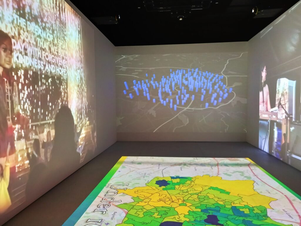 The digital exhibition had cameras projecting changing images onto long walls to the left and right and onto a shorter wall ahead, as well as onto the floor. The longer walls contain photographs. the shorter wall and floor feature maps of Coventry.