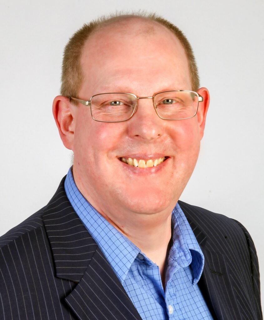 Head and shoulder image of Tim Hammerton. He has receding brown hair and wears golden-framed glasses. He is wearing a blue shirt and a blue pin-striped jacket.