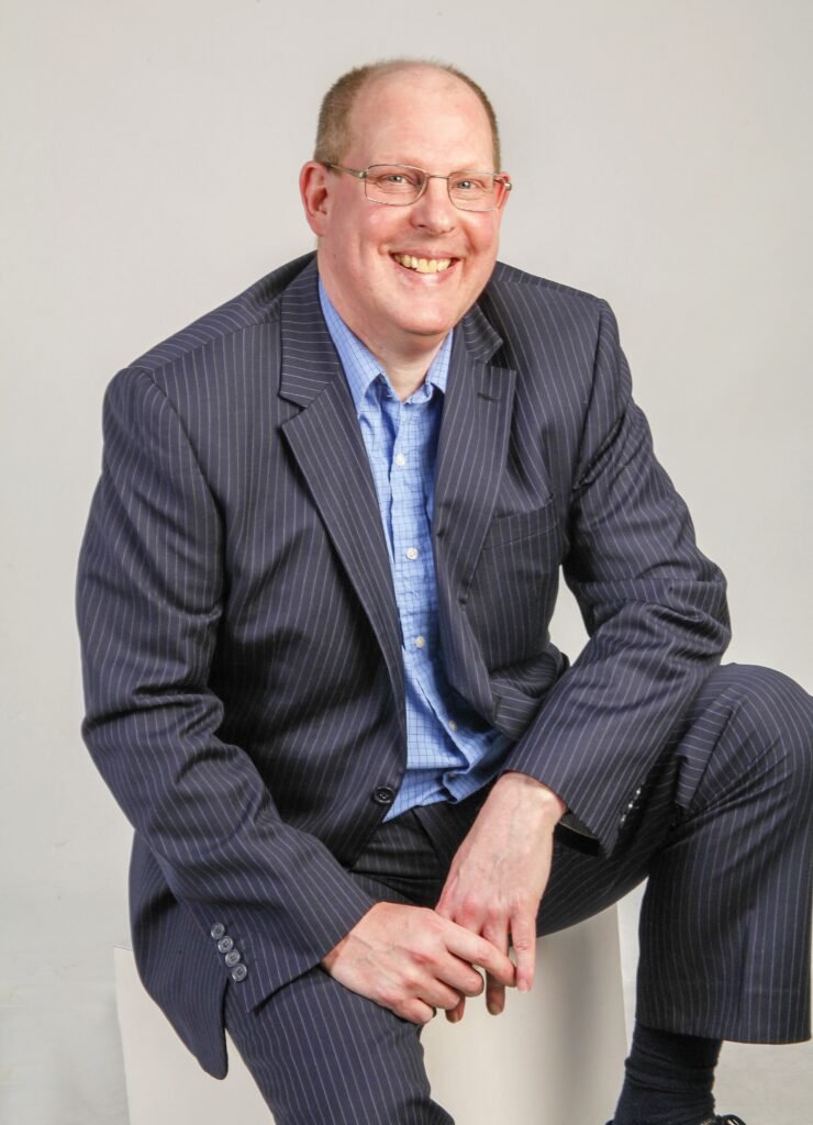 A full-length image of Tim Hammerton sitting down. He is smiling, has receding brown hair and wears golden-framed glasses. He is wearing a blue shirt and a blue pin-striped suit.
