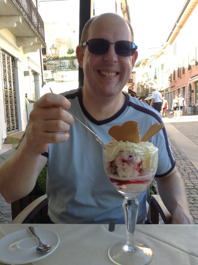 Tim Hammerton wearing sunglasses and a sky blue t-shirt. In front of him is a ice cream in a long glass. He is smiling, as he is about to tuck in using a long-handled spoon.