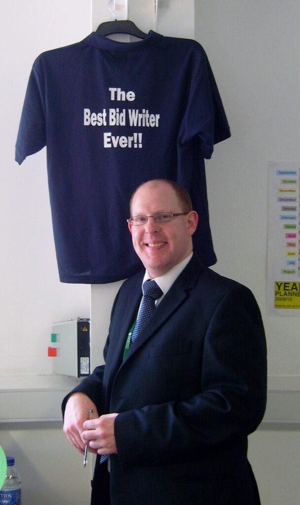 A photograph of Tim Hammerton, in suit and tie, standing below a t-shite hanging on the wall. The words on the t-shirt are 'The Best Bid Writer Ever!!' due to my role writing bids and tenders to win business.